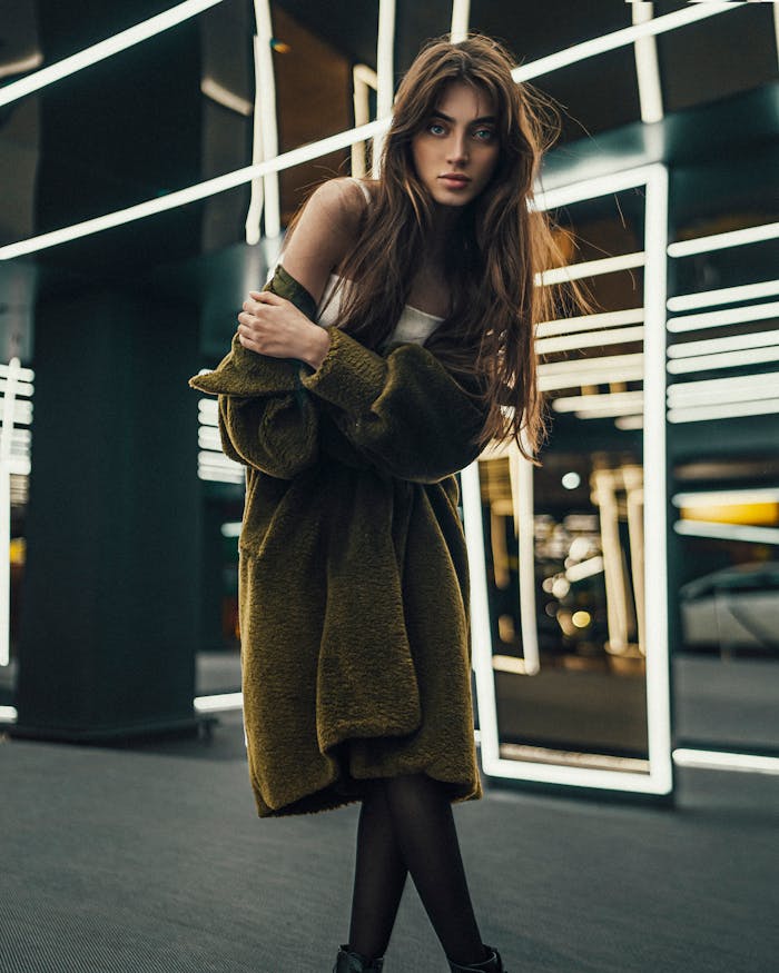 Chic young woman in green coat posing in a modern interior with illuminated mirrors in Istanbul.