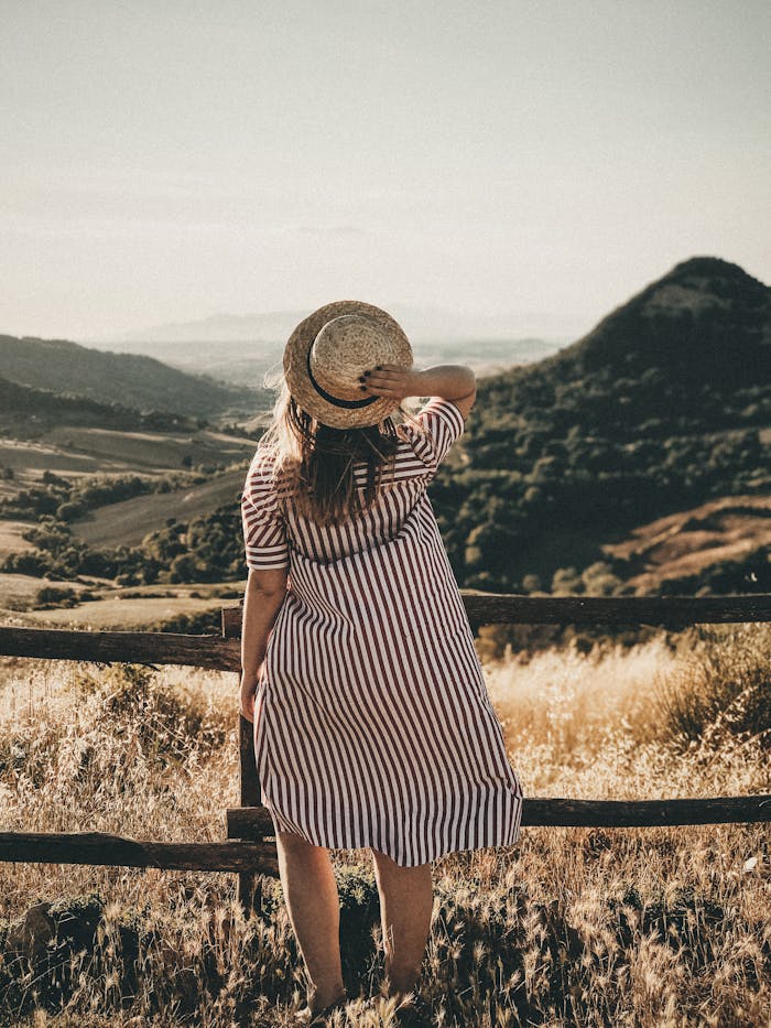 Young woman in striped dress and hat embracing a scenic countryside landscape during daylight.