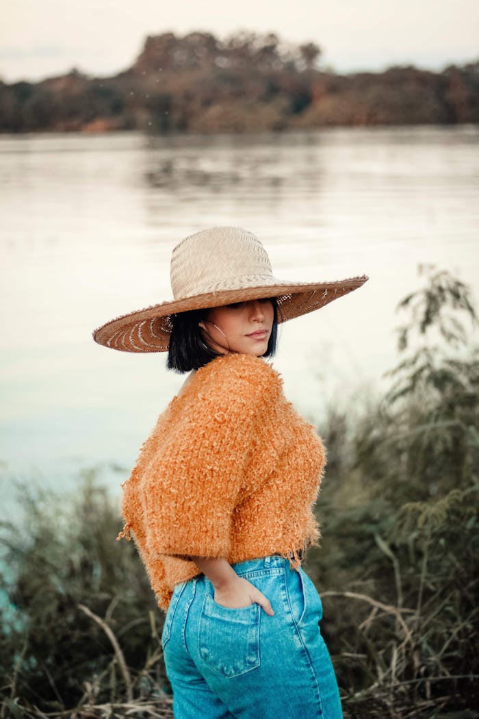 Stylish woman in denim shorts and a straw hat poses by the lake, showcasing casual fashion.
